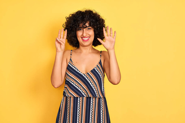 Mulher árabe com cabelo encaracolado vestindo vestido colorido listrado sobre fundo amarelo isolado mostrando e apontando para cima com os dedos número oito enquanto sorri confiante e feliz
. - Foto, Imagem