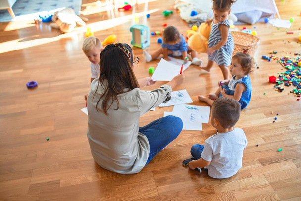 Красивий вчитель і група малюків, які сидять на підлозі малюють за допомогою паперу і олівця навколо безлічі іграшок в дитячому саду
 - Фото, зображення