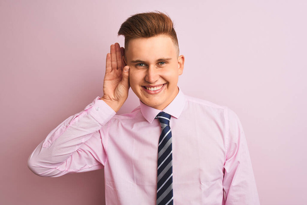 シャツとネクタイを着た若いハンサムなビジネスマンは、噂やゴシップに耳を傾けて耳を上げて微笑む孤立したピンクの背景の上に立っています。聴覚障害の概念. - 写真・画像