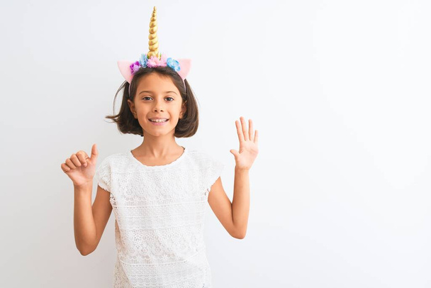 mooi kind meisje dragen eenhoorn diadeem staande over geïsoleerde witte achtergrond tonen en wijzen met vingers nummer zes terwijl glimlachen zelfverzekerd en gelukkig. - Foto, afbeelding