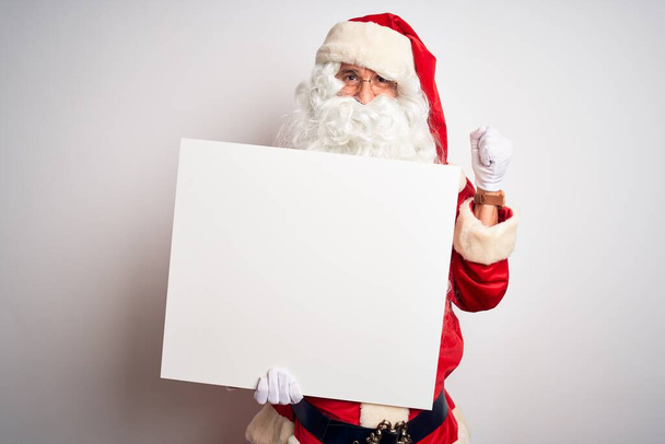 Noel Baba kostümü giymiş orta yaşlı bir adam beyaz arka planda afiş tutuyor gururla bağırıyor, zaferi kutluyor ve başarıyı kutluyor çok heyecanlı, coşkulu bir duygu. - Fotoğraf, Görsel
