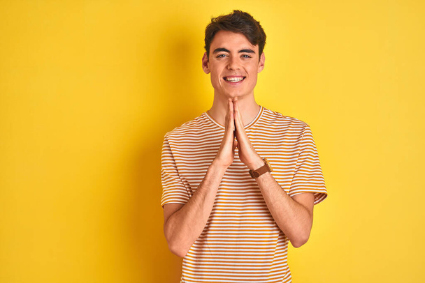 tiener jongen dragen geel t-shirt over geïsoleerde achtergrond bidden met de handen bij elkaar vragen om vergeving glimlachen zelfverzekerd. - Foto, afbeelding