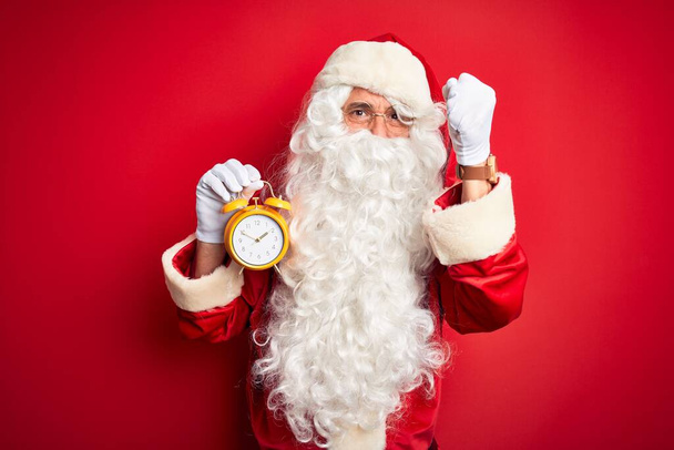 Ανώτερος άνθρωπος φορώντας κοστούμι Άγιος Βασίλης κρατώντας το ξυπνητήρι πάνω από απομονωμένο κόκκινο φόντο ενοχλημένος και απογοητευμένος φωνάζοντας με θυμό, τρελός και φωνάζοντας με σηκωμένο χέρι, έννοια του θυμού - Φωτογραφία, εικόνα