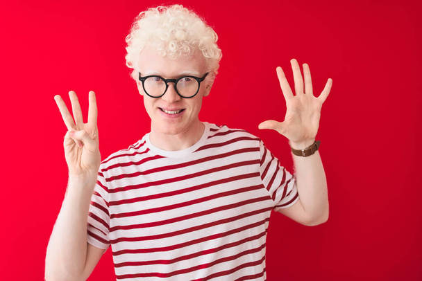 Νέοι Albino ξανθός άνδρας φορώντας ριγέ t-shirt και γυαλιά πάνω από απομονωμένο κόκκινο φόντο δείχνει και δείχνει προς τα πάνω με τα δάχτυλα αριθμό οκτώ, ενώ χαμογελά αυτοπεποίθηση και χαρούμενος. - Φωτογραφία, εικόνα