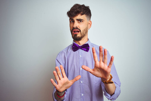 Mladý muž s tetováním v purpurové košili a motýlku na izolovaném bílém pozadí, vyděšený a vyděšený výrazem strachu, se zastaví a vykřikne v šoku. Panický koncept. - Fotografie, Obrázek