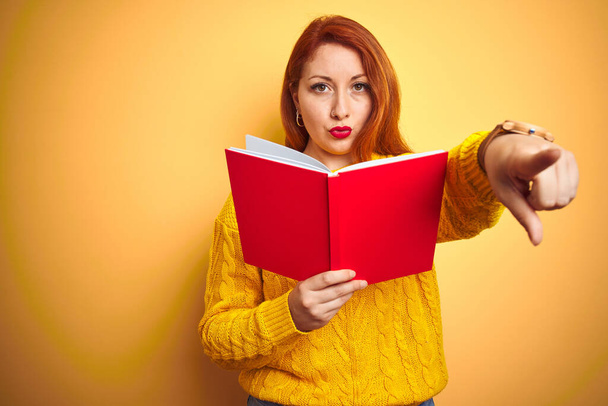 Молодая рыжая учительница читает красную книгу на желтом изолированном фоне, указывая пальцем на камеру и на вас, знак руки, позитивный и уверенный жест спереди
 - Фото, изображение
