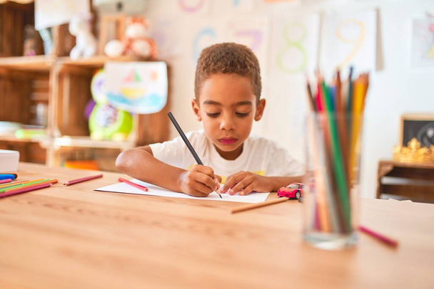 Hermoso niño afroamericano sentado dibujo usando papel y lápices en el escritorio en el jardín de infantes
 - Foto, Imagen
