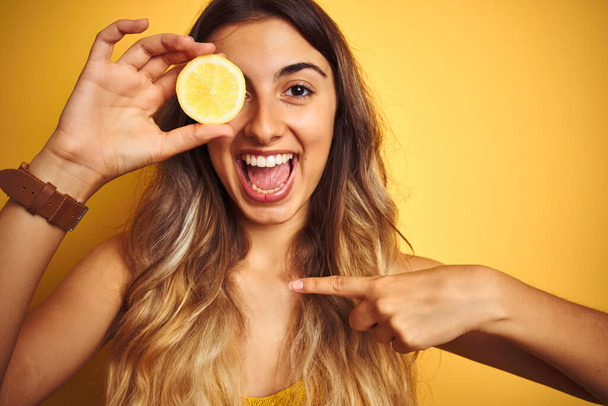 Nuori kaunis nainen, jolla on sitruuna silmässä keltaisen taustan päällä, yllättäen kasvot osoittaen sormella itseään
 - Valokuva, kuva