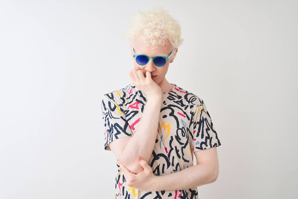 Mladý albín blond muž nosí barevné tričko a sluneční brýle přes izolované červené pozadí myšlení vypadá unaveně a znuděný deprese problémy se zkříženými pažemi. - Fotografie, Obrázek