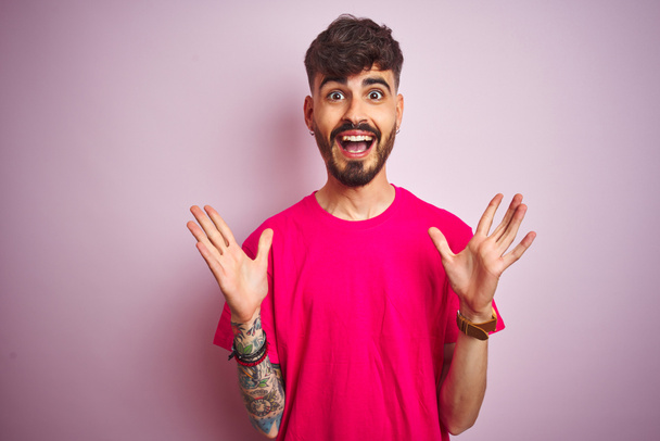 Νεαρός άνδρας με τατουάζ φορώντας t-shirt στέκεται πάνω από απομονωμένο ροζ φόντο γιορτάζει τρελό και έκπληκτος για την επιτυχία με τα χέρια υψωμένα και ανοιχτά μάτια ουρλιάζοντας ενθουσιασμένος. Έννοια νικητή - Φωτογραφία, εικόνα