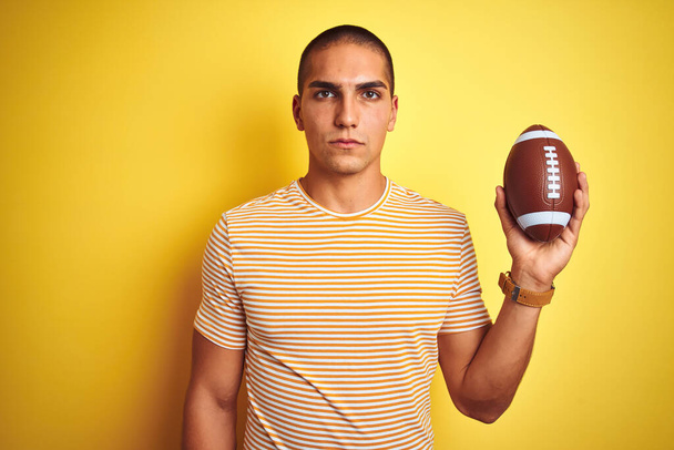 Νεαρός παίκτης ράγκμπι κρατώντας μια μπάλα ποδοσφαίρου πάνω από κίτρινο απομονωμένο φόντο με μια σίγουρη έκφραση στο έξυπνο πρόσωπο σκέψης σοβαρή - Φωτογραφία, εικόνα