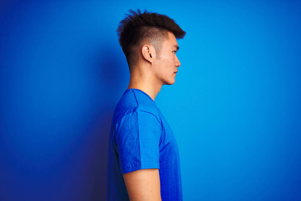 Молодой азиатский китайский мужчина в футболке стоя на изолированном синем фоне глядя в сторону, расслабиться профиль позировать с естественным лицом с уверенной улыбкой
. - Фото, изображение
