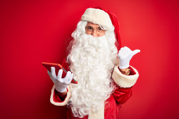 Μεσήλικας άνδρας φορώντας κοστούμι Άγιος Βασίλης χρησιμοποιώντας tablet πάνω από απομονωμένο κόκκινο φόντο που δείχνει και δείχνει με τον αντίχειρα μέχρι το πλάι με χαρούμενο πρόσωπο χαμογελώντας - Φωτογραφία, εικόνα