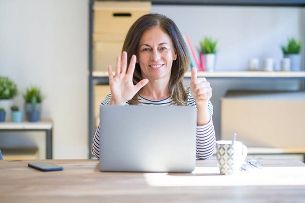 自信と幸せ笑顔ながらコンピュータのラップトップを使用して自宅で仕事でテーブルに座っている中年のシニア女性は指の数6を示すと指摘します. - 写真・画像