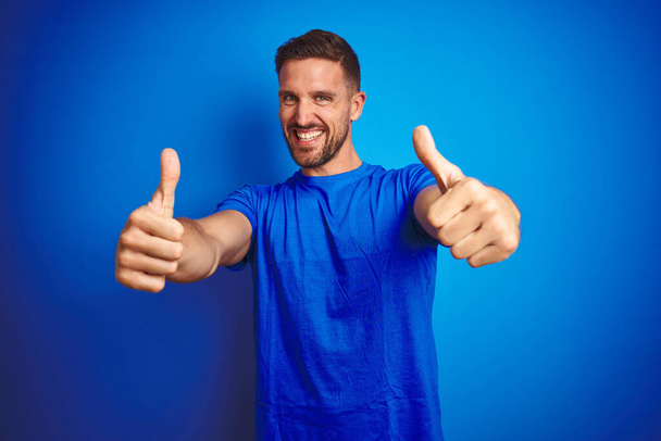 Young handsome man wearing casual t-shirt over blue isolated background approving doing positive gesture with hand, thumbs up smiling and happy for success. Winner gesture. - Photo, Image