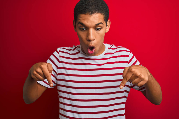 Młody przystojny arabski mężczyzna w pasiastej koszulce na izolowanym czerwonym tle wskazując palcami pokazującymi reklamę, zaskoczoną twarz i otwarte usta - Zdjęcie, obraz