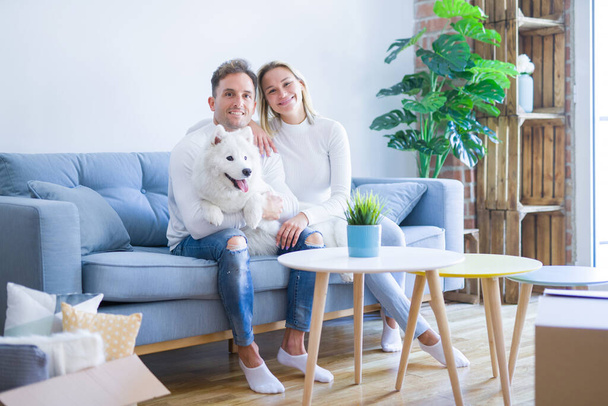 Young beautiful couple with dog sitting on the sofa at new home around cardboard boxes - Fotografie, Obrázek