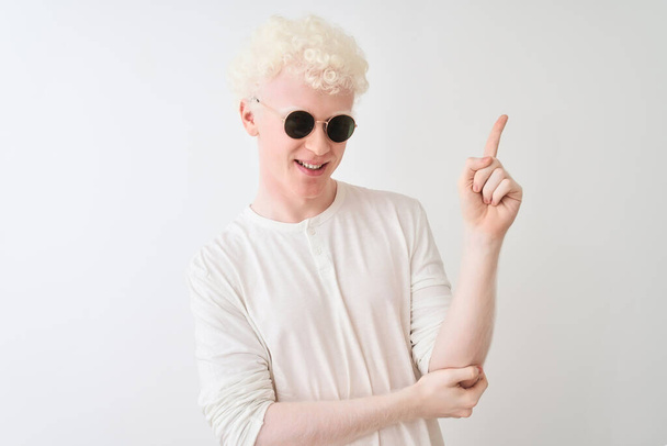 Młody blondyn albinos w koszulce i okularach przeciwsłonecznych na białym tle z dużym uśmiechem na twarzy, wskazując ręką i palcem w bok patrząc w kamerę. - Zdjęcie, obraz