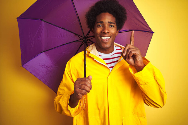 雨のコートと傘を身に着けているアフリカ系アメリカ人男性は、幸せな顔をしたアイデアや質問指差し指で驚いて孤立した黄色の背景の上に立って、ナンバーワン - 写真・画像