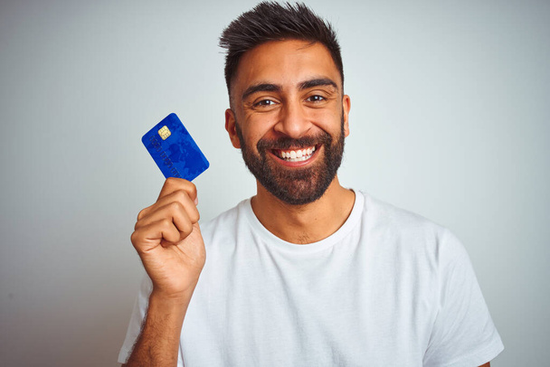 Mladý indický zákazník muž držící kreditní kartu stojící nad izolovaným bílým pozadím s veselou tváří stojící a usmívající se s sebevědomým úsměvem ukazující zuby - Fotografie, Obrázek