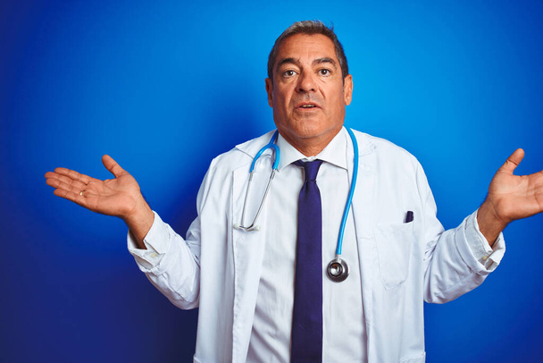 Handsome middle age doctor man wearing stethoscope over isolated blue background clueless and confused expression with arms and hands raised. Doubt concept. - Фото, изображение