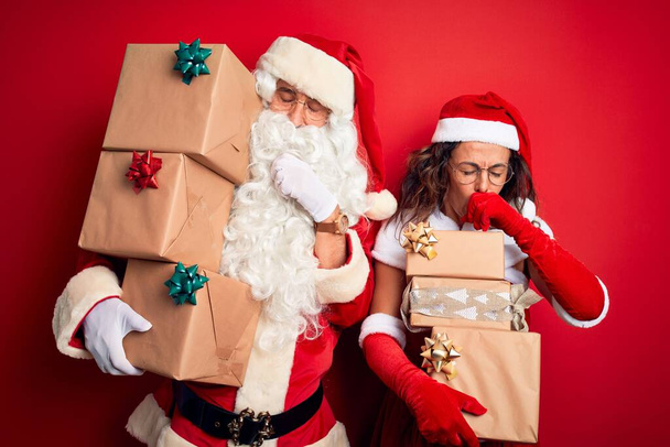Pár středního věku v kostýmu Santa drží věž darů na izolovaném červeném pozadí pocit nevolnosti a kašle jako symptom pro nachlazení nebo bronchitidy. Koncept zdravotní péče. - Fotografie, Obrázek