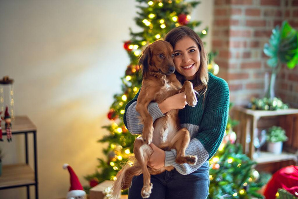 Young beautiful woman smiling happy and confident. Holding and hugging dog around christmas tree at home - Photo, Image