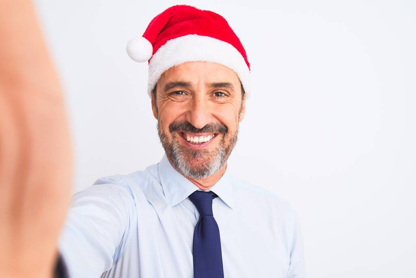Middelbare leeftijd zakenman het dragen van kerst hoed maken selfie over geïsoleerde witte achtergrond met een gelukkig gezicht staande en glimlachend met een zelfverzekerde glimlach tonen tanden - Foto, afbeelding