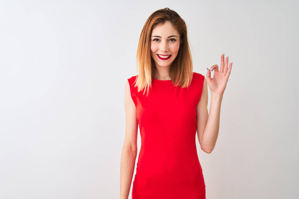 Empresária ruiva vestindo elegante vestido vermelho em pé sobre fundo branco isolado sorrindo positivo fazendo sinal ok com a mão e os dedos. Expressão bem sucedida
. - Foto, Imagem