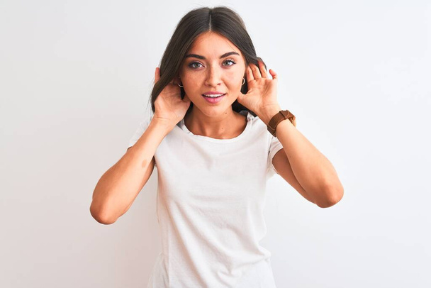 Jonge mooie vrouw draagt casual t-shirt staan over geïsoleerde witte achtergrond Proberen om beide handen te horen op oor gebaar, nieuwsgierig naar roddels. Gehoorproblemen, doof - Foto, afbeelding