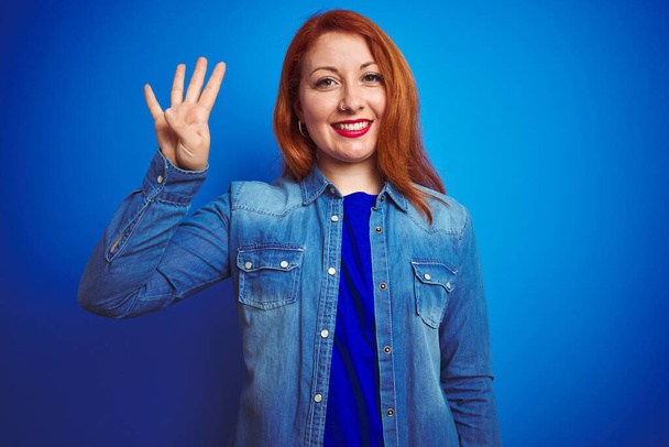 Mavi izole edilmiş arka plan üzerinde duran güzel kızıl saçlı kadın dört numaralı parmaklarıyla gülümseyerek kendinden emin ve mutlu bir şekilde işaret ediyor.. - Fotoğraf, Görsel