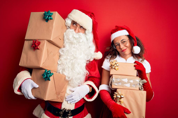 Couple d'âge moyen portant costume de Père Noël tenant tour de cadeaux sur fond rouge isolé regardant somnolent et fatigué, épuisé par la fatigue et la gueule de bois, les yeux paresseux le matin
. - Photo, image