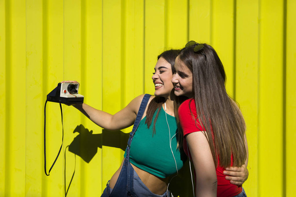 Dos mujeres felices bastante jóvenes tomando fotos con cámara retro y pasar un buen rato por la pared amarilla
 - Foto, imagen