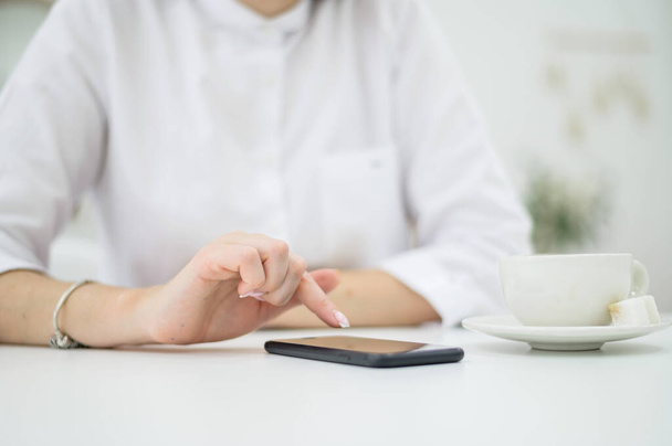 A woman in a white shirt with well-groomed hands and fresh manicure is drinking coffee in a cafe. Girl writes a message on a smartphone at lunchtime. Close-up - Photo, Image