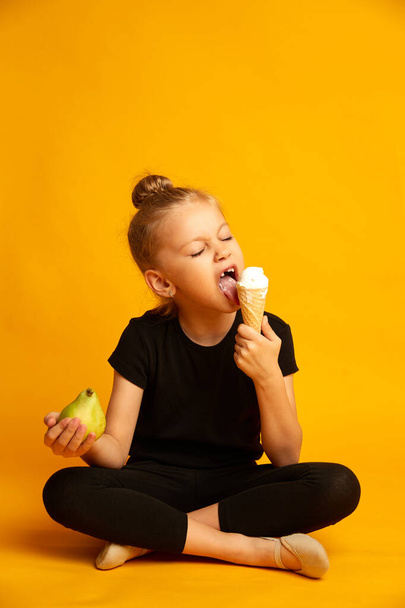 Cute little dancer girl choosing between apple and sweet ice-cream on yellow background - Fotó, kép