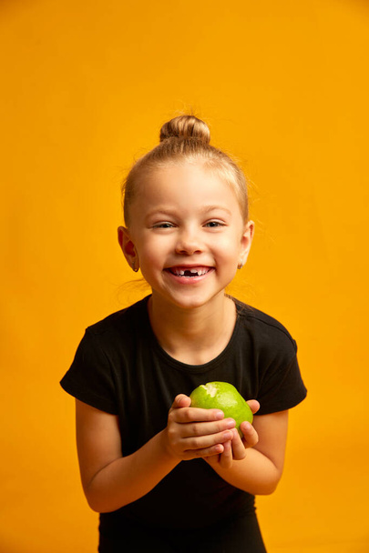 little dancer portrait on yellow background, portrait of a happy child smiling and showing off her first lost milk tooth in her hand a green apple - Photo, Image