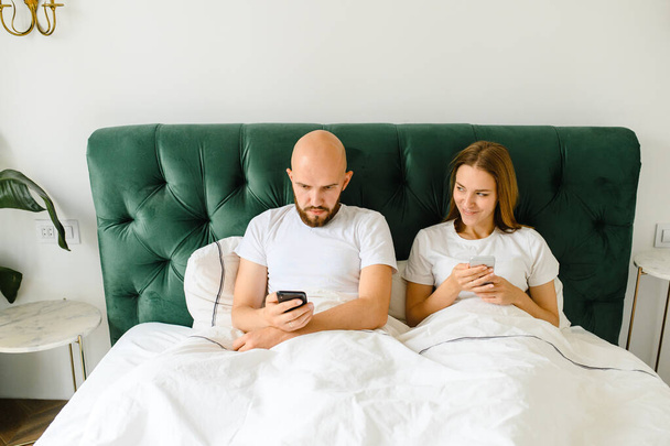 young couple lie in bed with phones. Communication and relationship problems. - Zdjęcie, obraz