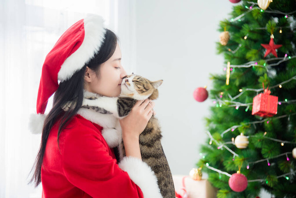 schöne asiatische Mädchen im Weihnachtsmann-Kostüm küsst ihre Katze im Wohnzimmer - Foto, Bild