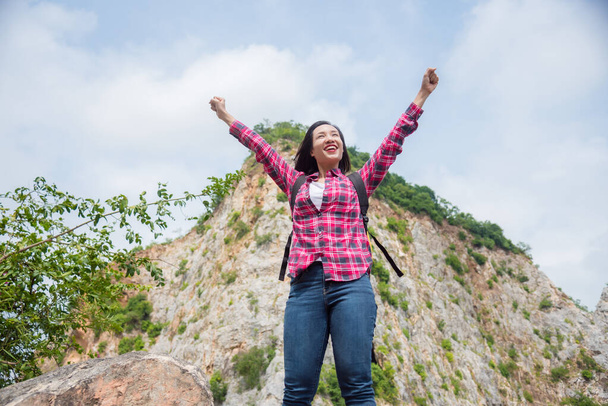 feliz asiático viajante mulher com mochila de pé no pedra colina e sorrisos com levantando as mãos
. - Foto, Imagem