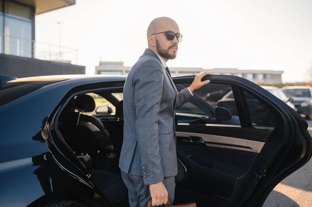 Portrait of a handsome businessman in sunglasses standing near the car outdoors in front of the modern building facade - Foto, afbeelding