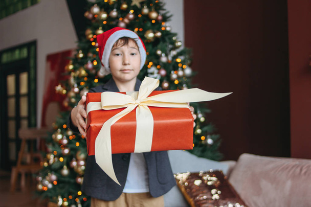 Attractive funny tween boy with dark hair in santa hat gives present at camera on christmas tree background - 写真・画像