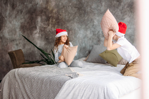 niedlich zwischen Kindern in Weihnachtsmützen und Pyjamas, die auf dem Bett mit Kissen spielen, Weihnachten morgens, Kinderparty - Foto, Bild