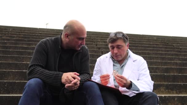 Médico examinando um relatório médico sentado em escadas ao ar livre conversando com um paciente e explicando os resultados
 - Filmagem, Vídeo