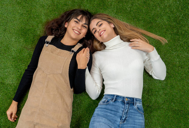 Top view of two stylish girl friends having fun laughing and making faces lying down on grass. Blonde and brunette happy young teenagers women enjoying time together. Friendship, love and care. - Photo, image