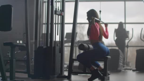 Una joven está entrenando su espalda en el gimnasio. Entrenamiento en una habitación con grandes ventanas en el fondo una mujer utiliza un elipsoide y una cinta de correr
 - Metraje, vídeo