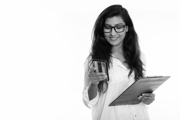 Studio shot of young happy Persian woman smiling while using mobile phone and holding clipboard - Foto, Imagen