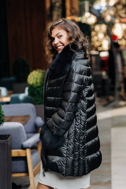 Charming girl dressed in black down jacket with fur poses in the street - Photo, Image