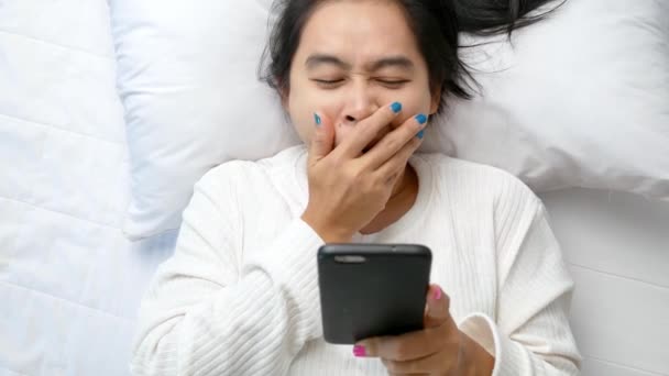 Asian woman using smartphone and yawning while lying on bed at home. Technology and relaxation concept. - Filmati, video
