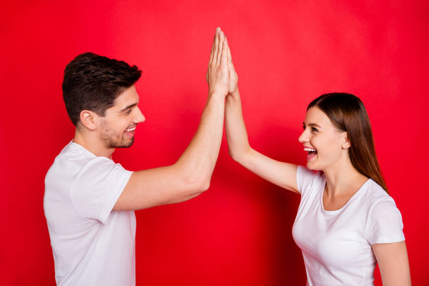 Photo of cheerful cute nice positive pretty boyfriend students girlfriend clapping their palms giving high five smiling toothily laughing in white t-shirts isolated vivid red color background - 写真・画像