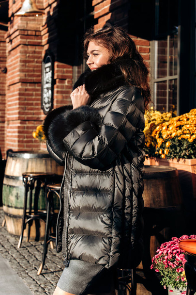 Charming girl dressed in black down jacket with fur poses in the street - Фото, зображення